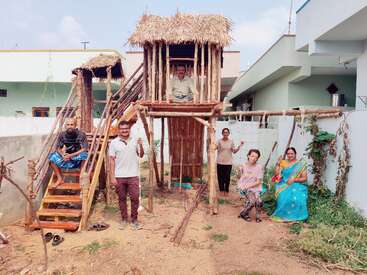 Sechs Personen posieren um ein handgefertigtes hölzernes Baumhaus mit Strohdach, Schaukeln und Treppen in einem Hinterhof, umgeben von Grünflächen und modernen Wohngebäuden.