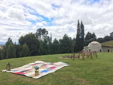 The image depicts a serene outdoor setting with a vibrant quilted blanket, a lantern, and a vase of flowers on a lush grassy hill, surrounded by trees and a cloudy sky.