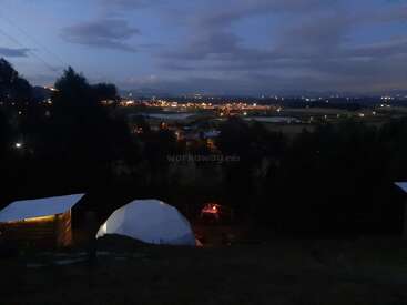The image depicts a serene nighttime scene featuring a tent and a building with lights on, set against a backdrop of a town and a cloudy sky.