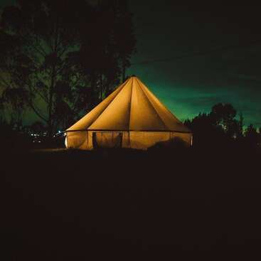 The image depicts a large tent illuminated from within at night, set against a dark background with trees visible in the distance.