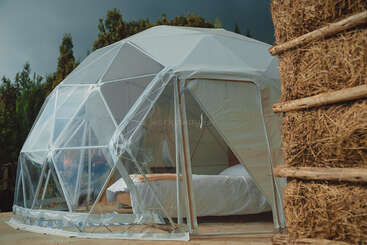 A modern geodesic dome tent with transparent panels sits outdoors. Inside, a bed is visible. Trees and hay bales surround the structure under a cloudy sky.
