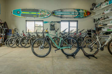 Esta imagem mostra uma garagem ou loja bem organizada, com várias bicicletas, duas pranchas de paddle coloridas montadas na parede e racks com vários equipamentos e acessórios para atividades ao ar livre.