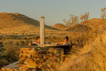 Duas pessoas relaxam em uma banheira de hidromassagem rústica ao ar livre construída com pedra, cercada por grama seca e colinas do deserto, desfrutando de bebidas e do sereno pôr do sol dourado.