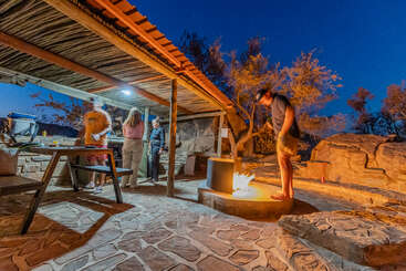 Quatro pessoas se reúnem ao ar livre ao anoitecer, sob uma pérgola rústica. Uma pessoa cuida de uma fogueira, enquanto as outras preparam a comida e socializam, aproveitando a atmosfera quente da noite.