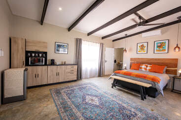 Este quarto aconchegante tem uma cama king-size com detalhes em laranja, móveis modernos de madeira, cozinha compacta, tapete colorido, vigas expostas, luzes pendentes e arte emoldurada nas paredes.