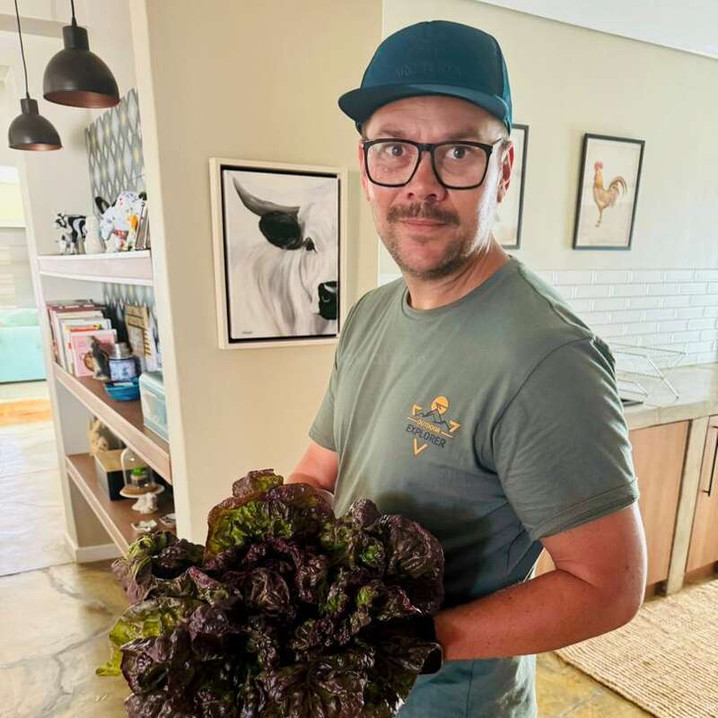 Um homem usando óculos e boné azul está em um ambiente interno, segurando folhas verdes frescas. Você pode ver a cozinha e a arte ao fundo, com prateleiras e quadros de animais emoldurados.