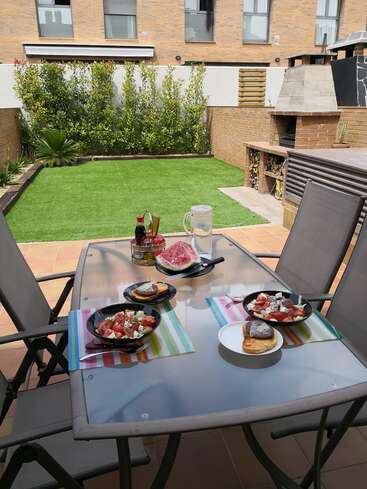 The image depicts a serene outdoor patio scene, featuring a table set with food and drinks, surrounded by a lush green lawn and a brick building in the background.
