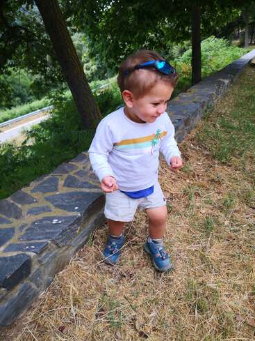 The image depicts a young boy standing on a stone wall, wearing a white shirt with a palm tree design, shorts, and blue shoes, in a grassy area with trees and a road in the background.