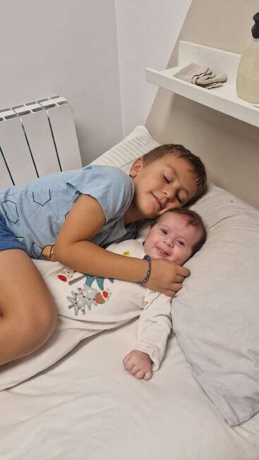 A young boy lovingly cuddles his baby sibling on a bed. The boy smiles with his eyes closed, while the baby looks happy and comfortable. Cozy moment.