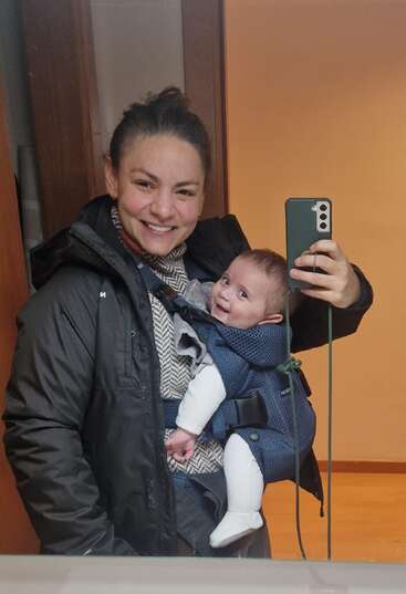 A smiling woman takes a mirror selfie, wearing a black jacket and scarf. She carries a happy baby in a blue front carrier, both looking cheerful.