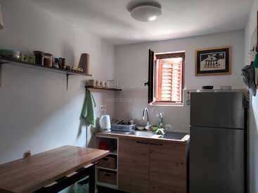 Eine einfache, kleine Küche mit Holzschränken, offenen Regalen, einem Kühlschrank, Arbeitsplattengeräten, einem Fenster mit Tageslicht und einem gerahmten Kunstwerk an der Wand.