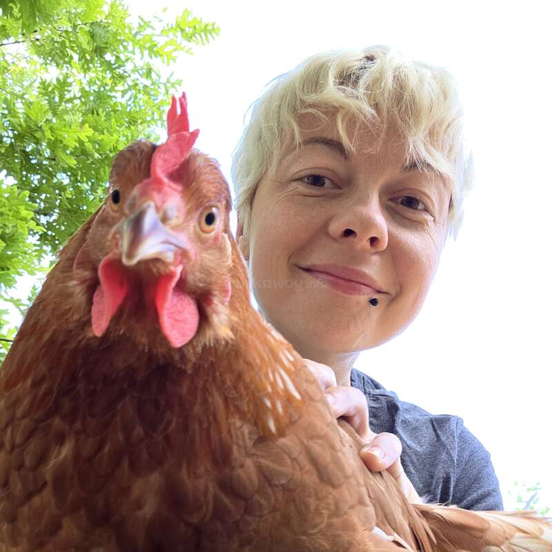 Eine lächelnde Person mit kurzen blonden Haaren hält ein braunes Huhn nahe an die Kamera. Oben sind grüne Blätter zu sehen, und der Himmel ist strahlend weiß.