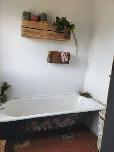 A cozy bathroom with a white bathtub, floral motifs, terracotta tiles, wooden crate shelves holding towels and a plant, and simple natural decorative elements.