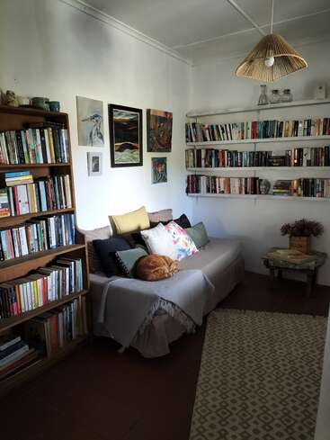 A cozy reading nook features bookshelves, a beige couch with colorful pillows, a sleeping orange cat, wall art, a small table, and a patterned rug.