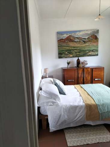 A cozy bedroom featuring a neatly made bed with white linens, light green and beige throws, wooden furniture, a landscape painting, and simple, minimalist decor.