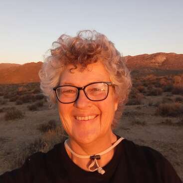Das Bild zeigt eine ältere Frau mit kurzen, lockigen Haaren und Brille, die bei Sonnenuntergang vor einer Wüstenlandschaft mit Bergen im Hintergrund lächelt.