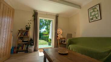Uma sala de estar aconchegante e bem iluminada, com um sofá verde, uma mesa de centro de madeira, estantes de livros, uma escrivaninha e uma porta que dá para um jardim verdejante e bem iluminado lá fora.