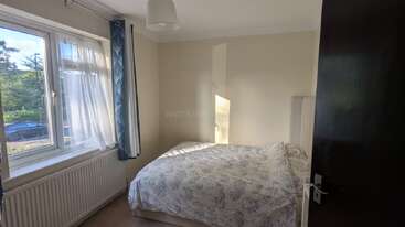 Este é um quarto pequeno e aconchegante com uma cama de casal, roupa de cama com estampa floral clara, uma janela grande com cortinas azuis, paredes em tom creme, luz natural e uma decoração simples.