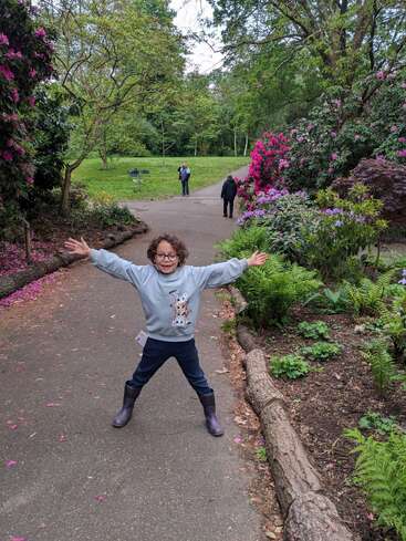 Uma criança alegre está em um caminho de jardim com os braços abertos, cercada por flores coloridas em plena floração, árvores verdes e um casal caminhando ao fundo.