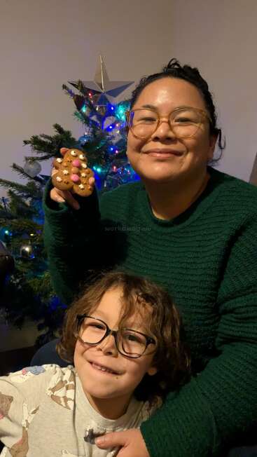 Uma mulher e uma criança sorriem em frente a uma árvore de Natal enfeitada. A mulher segura um biscoito de gengibre; ambas usam óculos e aproveitam juntas um momento festivo e aconchegante.
