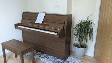 Um piano vertical marrom com um banco combinando está em um canto, sobre um tapete decorativo. Há partituras em cima do piano. Perto dali, há um vaso de planta.