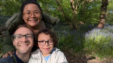 Uma família de três pessoas sorri alegremente para uma selfie em uma floresta exuberante, cercada por árvores verdes e flores roxas em flor sob um sol radiante.