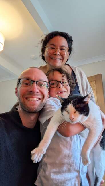 Três pessoas sorridentes e um gato preto e branco posam para uma foto alegre tirada dentro de casa. O gato, com os olhos fechados, está no centro, segurado com carinho.