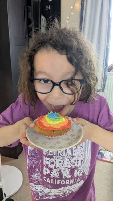 Uma criança pequena de óculos segura animadamente um prato com um cupcake colorido em tons de arco-íris, pronta para dar uma grande mordida, vestindo uma camiseta roxa da Califórnia.