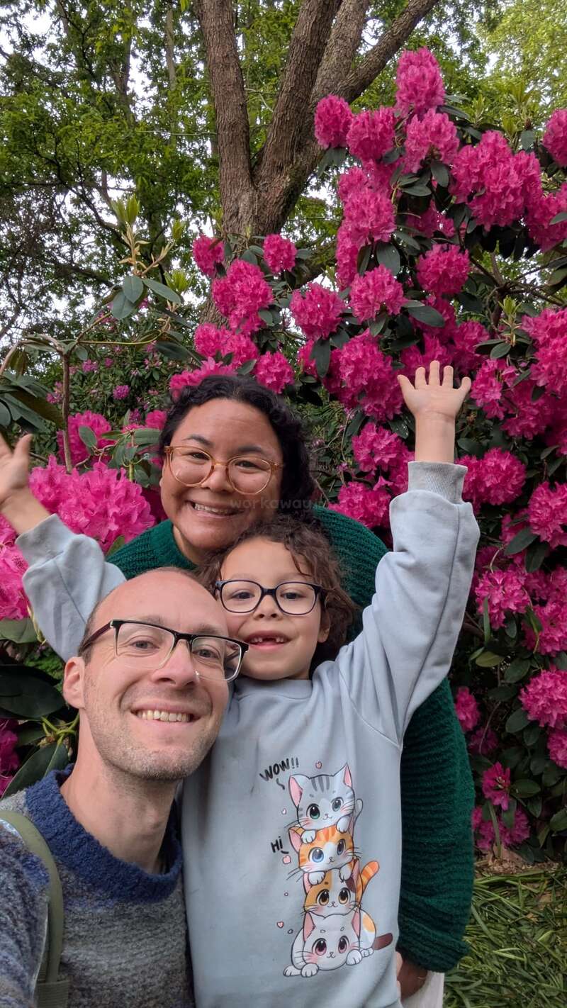 Uma alegre família de três pessoas sorri diante de flores rosa vibrantes. A criança levanta os braços com entusiasmo, enquanto todos usam óculos e parecem felizes ao ar livre.