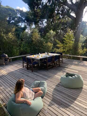 Uma mulher relaxa em um pufe em um deck de madeira cercado por árvores frondosas, com uma mesa de jantar e cadeiras preparadas para uma refeição ao ar livre.
