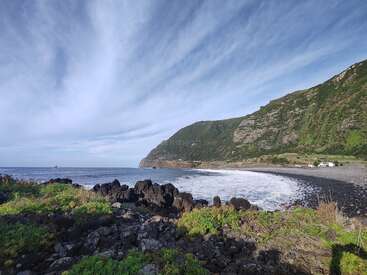 A beautiful coastal scene features rocky black shoreline, lush green cliffs, rolling waves, and a wide blue sky streaked with clouds, invoking tranquility and natural wonder.