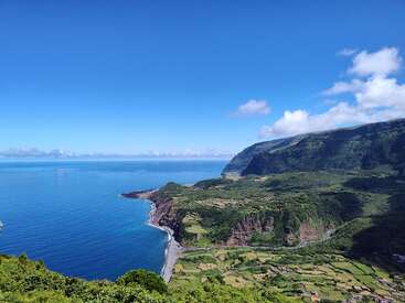 A breathtaking coastal landscape with lush green cliffs, terraced fields, a winding shoreline, and clear blue ocean under a bright sky dotted with scattered white clouds.