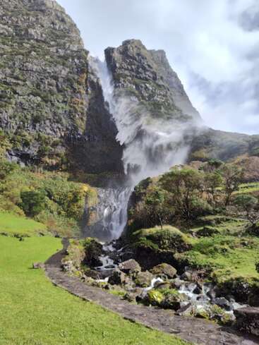 A stunning waterfall cascades down a steep rocky cliff, surrounded by lush greenery. A path winds through vibrant grass, leading towards the powerful, misty falls.