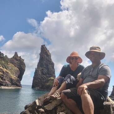 A smiling couple sits on rocks by the sea, wearing hats and sunglasses. Behind them, tall rocky sea stacks rise dramatically under a partly cloudy sky.