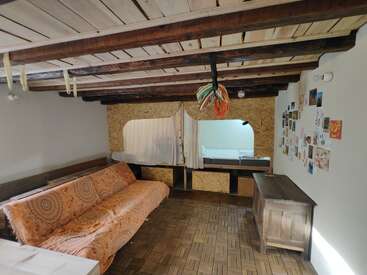 This cozy room features wooden beams, a patterned orange sofa, parquet flooring, a wall with photos, a trunk, and a curtained sleeping nook with a bed.