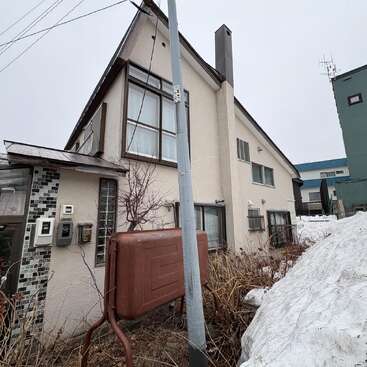 Uma casa bege com janelas grandes e telhado inclinado fica ao lado de um monte de neve, um poste elétrico, um tanque de metal e plantas crescidas, sob um céu cinzento e nublado.