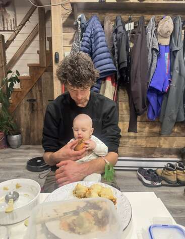 Un homme est assis avec un bébé sur les genoux et l'aide à manger. Des manteaux et des chaussures sont à l'arrière-plan et de la nourriture est posée sur la table.