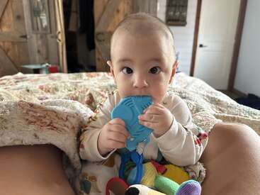 Un bébé aux grands yeux curieux est assis sur un lit, mâchant un jouet de dentition bleu, entouré de jouets colorés et enveloppé dans une couverture douillette.