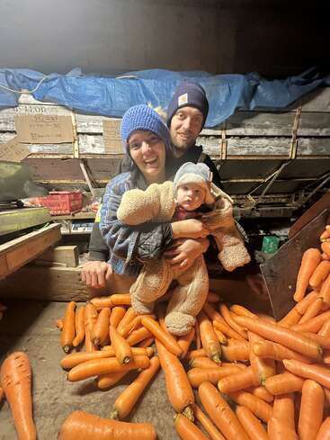 Une famille souriante de trois personnes, chaudement vêtues de bonnets et de vestes, pose joyeusement avec son bébé parmi des piles de grosses carottes fraîchement récoltées à l'intérieur d'une grange.