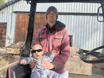 Un homme vêtu d'une veste à carreaux rouges et d'un bonnet se tient à l'extérieur, tenant un bébé portant des lunettes de soleil et un pyjama, à côté de caisses en bois et d'un bardage métallique. Journée ensoleillée.