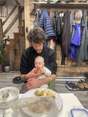Un homme est assis avec un bébé sur les genoux et l'aide à manger. Des manteaux sont accrochés au mur derrière, et de la nourriture est étalée sur la table. Un cadre confortable.