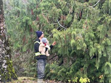 Un homme tenant un bébé se tient à côté d'arbres denses et verts, tous deux habillés chaudement. L'homme montre du doigt le feuillage, peut-être en train d'enseigner ou de partager quelque chose avec le bébé.