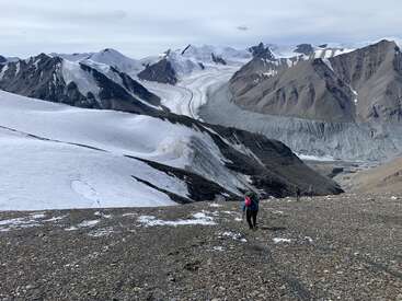 Trois randonneurs gravissent une pente rocheuse saupoudrée de neige. Des montagnes imposantes et escarpées ainsi que des glaciers s'étendent à perte de vue sous un ciel nuageux et immense. Une nature sauvage majestueuse les entoure.
