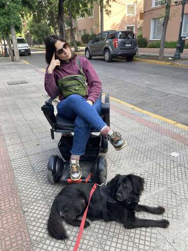 Una mujer con gafas de sol está sentada en una silla de ruedas eléctrica en la acera, con aire pensativo. Un perro negro atado con una correa roja yace tranquilamente a su lado.