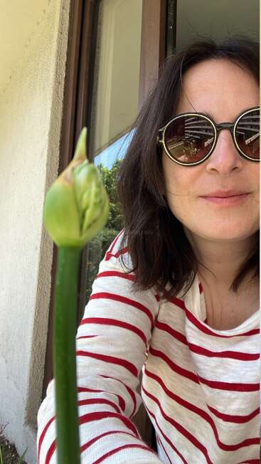 Una mujer con gafas de sol redondas y una camisa blanca a rayas rojas posa al aire libre junto a un capullo verde, cerca de una ventana, sonriendo bajo la luz natural del día.