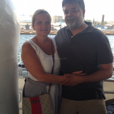 La imagen representa a una pareja abrazada en un barco, con una serena masa de agua y un muelle de fondo, evocando una sensación de tranquilidad y conexión.