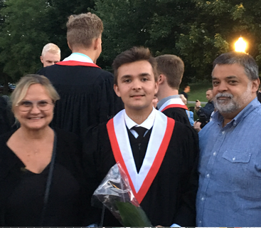 La imagen muestra a un joven vestido con un traje de graduación y una estola roja y blanca, rodeado de un grupo de personas en un entorno al aire libre.