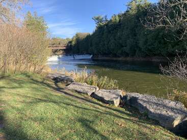 Ein ruhiger Fluss fließt unter einer Brücke hindurch, mit einem kleinen Wasserfall in der Ferne. Üppig grüne Bäume und grasbewachsene Ufer rahmen die friedliche Naturszene ein.