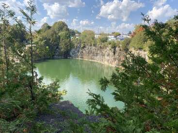 Ein ruhiger grüner See ist von felsigen Klippen und üppigen Bäumen umgeben. Der blaue Himmel mit flauschigen Wolken schafft eine friedliche, natürliche Szene, die von Laub umrahmt wird.