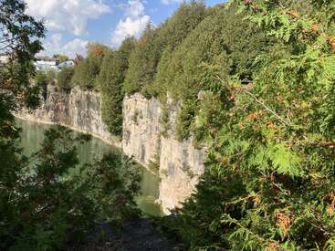 Eine felsige Klippe steht hoch neben einem grünlichen Fluss, umgeben von dichten immergrünen Bäumen unter einem strahlend blauen Himmel mit vereinzelten weißen Wolken. Friedliche Natur.